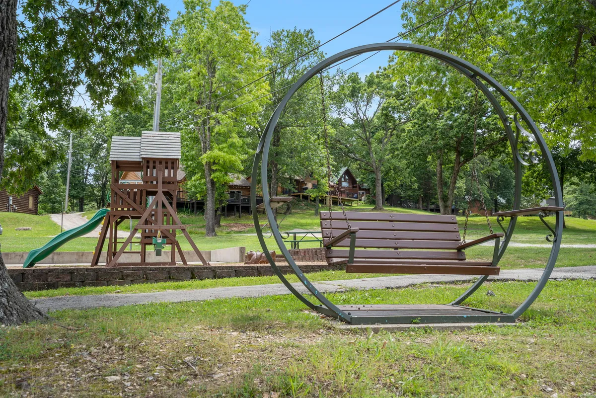 Children are laughing and playing on a well-equipped playground with a swing in a circular frame and a slide, while families connect at nearby picnic tables. Cozy cabins dot the background, adding to the communal, joyful vibe.