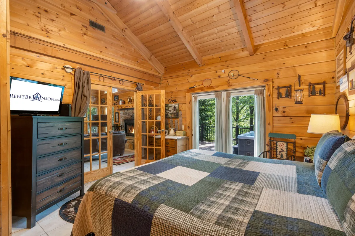 A cozy bedroom with a queen-size bed adorned with a plaid bedspread, surrounded by warm wooden walls. Large windows bring in natural light and offer woodland views. A TV is placed on a dresser, and the overall ambiance is restful and inviting.