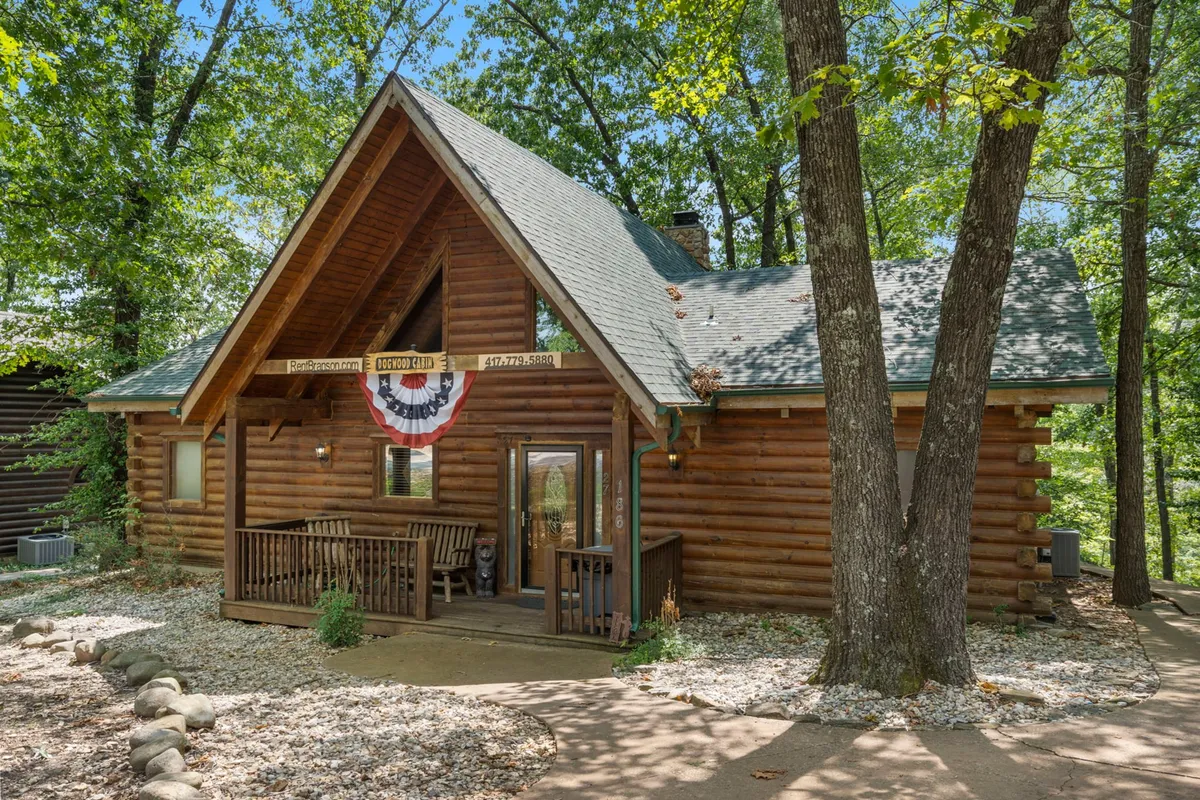 A charming log cabin set among leafy trees, with a welcoming front porch adorned with decorative bunting. The porch features comfortable seating and overlooks the natural wooded surroundings, evoking a sense of seclusion and retreat.