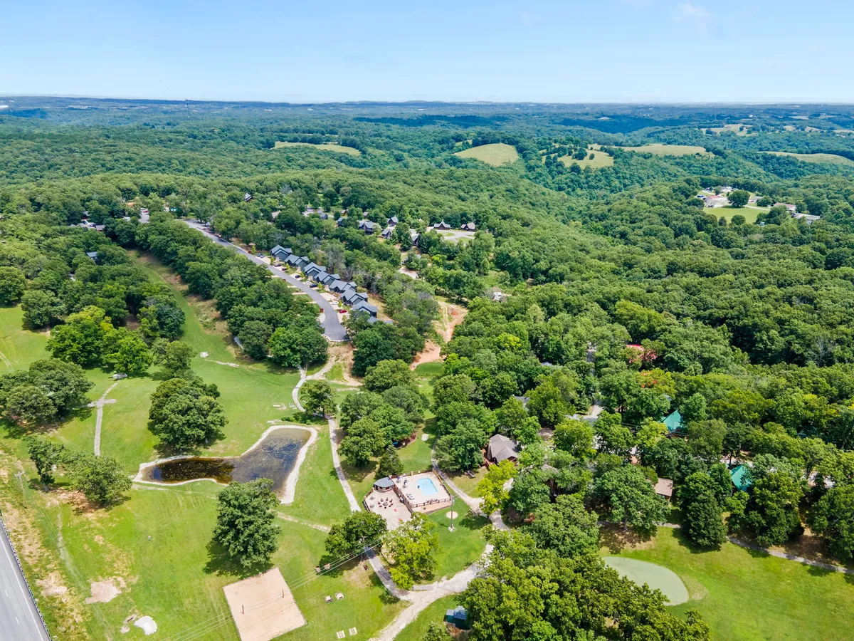 A wide view of the resort grounds, including an expansive green landscape, a small pond, swimming pool area, and a cluster of cabins connected by walking paths. The lush surroundings and open sky create a sense of adventure and exploration.