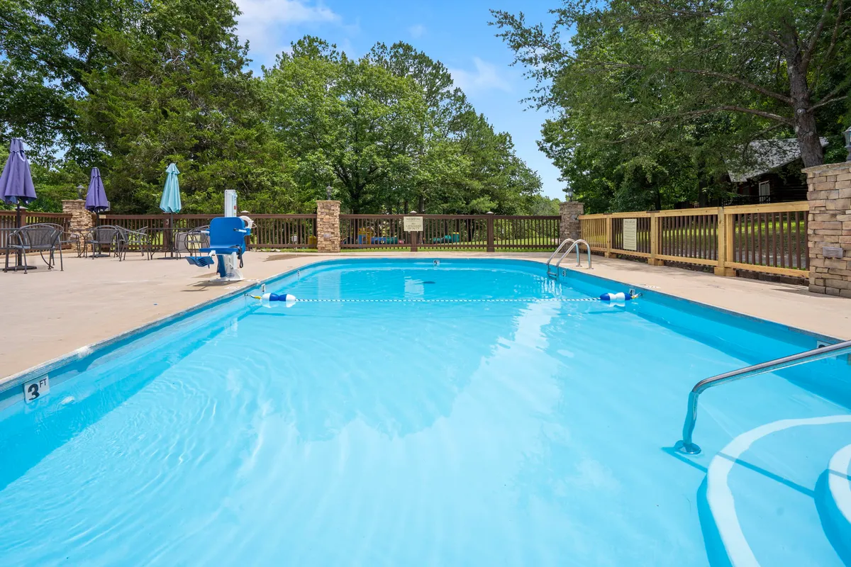 A sparkling outdoor swimming pool is surrounded by lush trees and plenty of lounge chairs, with umbrellas providing shade. The area is lively, with families and friends enjoying the water, socializing by the poolside, and relaxing under the sun.