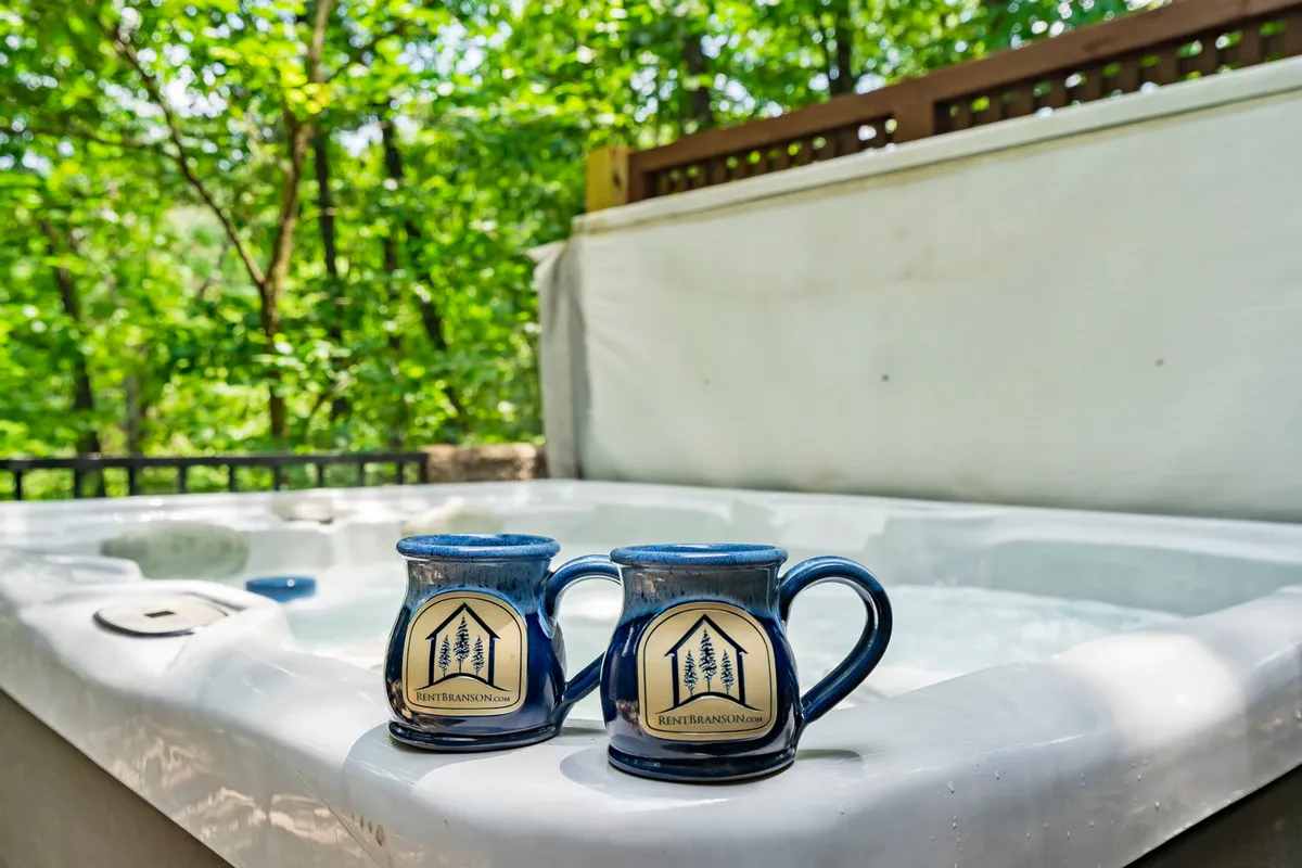 A private hot tub set on a wooden deck, surrounded by lush greenery. Two ceramic mugs rest on the tub’s edge, emphasizing relaxation and a connection to nature. The environment is tranquil and intimate, suggesting privacy and immersion in the outdoors.