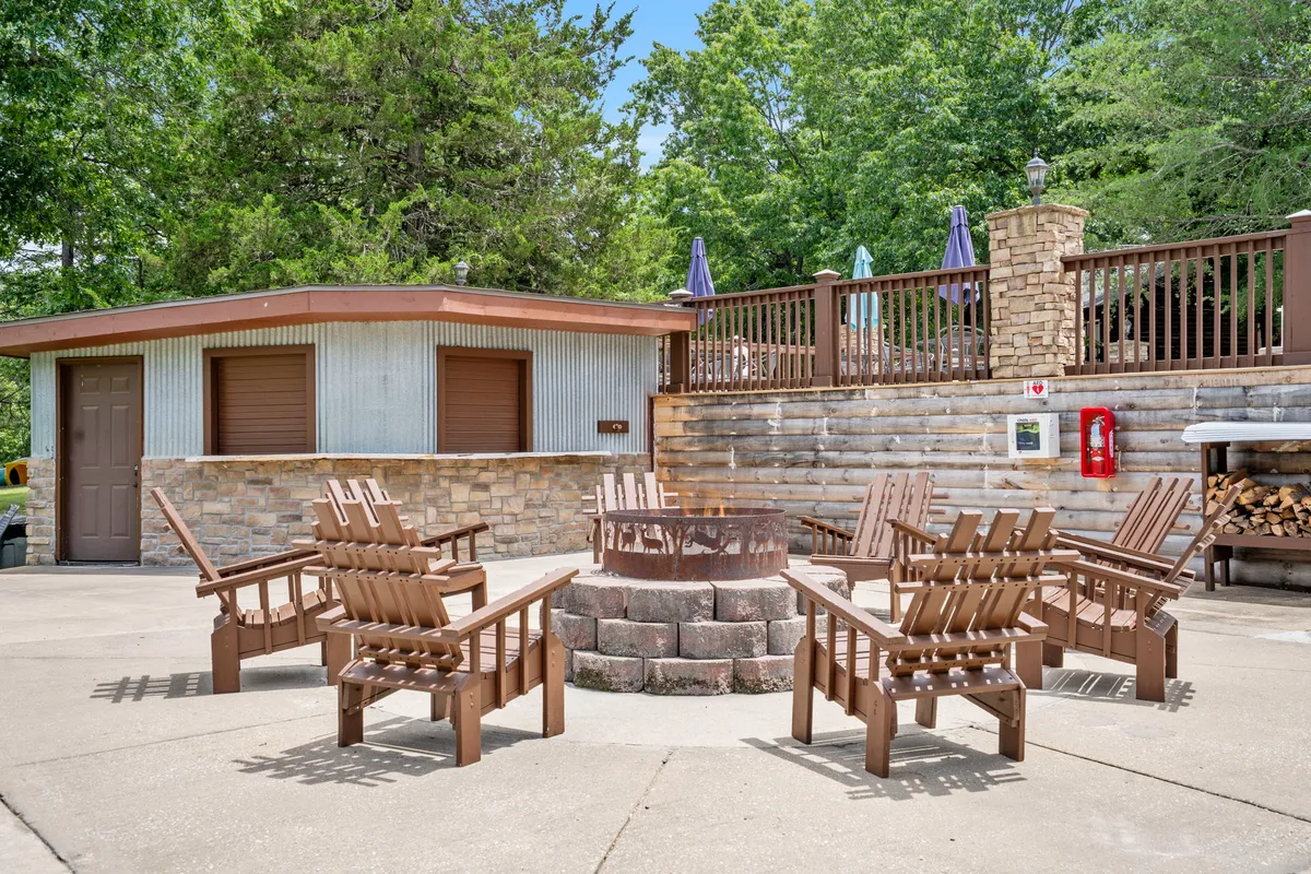 An exterior fire pit area with Adirondack chairs set in a circle, surrounded by lush greenery and a rustic building. The scene suggests a welcoming space for evening gatherings, storytelling, and toasting marshmallows under the stars.