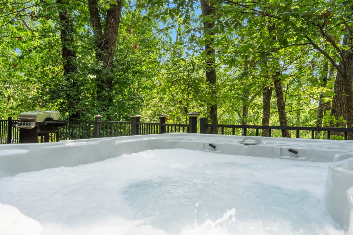 The image shows a charming log cabin nestled at the edge of lush woods, with a private fenced hot tub as the focal point. The surrounding greenery creates a sense of seclusion and nature, while the cozy outdoor space invites guests to unwind and connect. The scene is serene, suggesting opportunities for sunset soaks, morning coffee in the fresh air, and peaceful evenings under the stars.