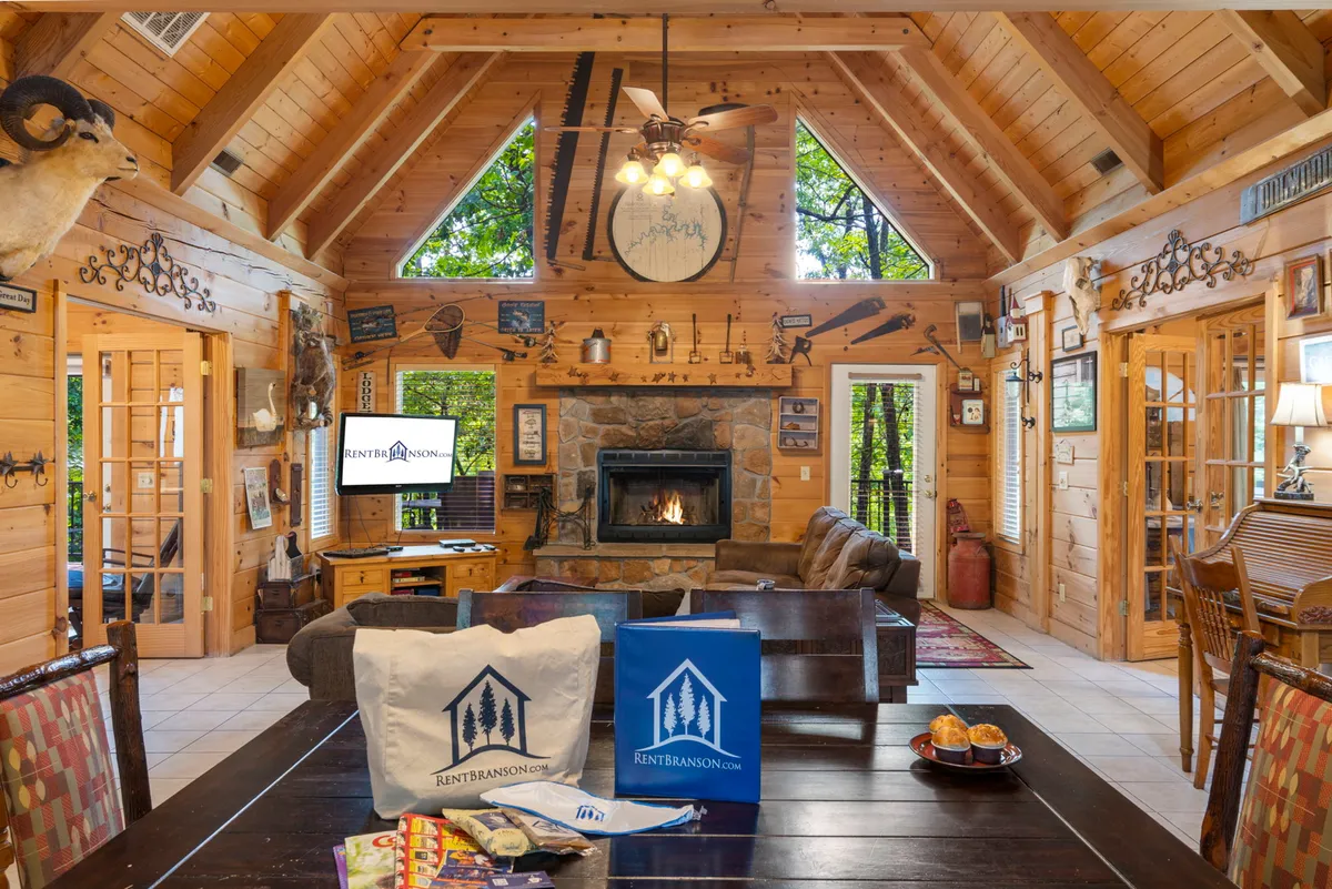 This interior scene showcases a spacious great room with a prominent stone fireplace, exposed wooden beams, large windows bringing in plenty of natural light, and a dining table set for gathering. The arrangement hints at a comfortable, welcoming environment ideal for family gatherings, cozy evenings, and celebrations.