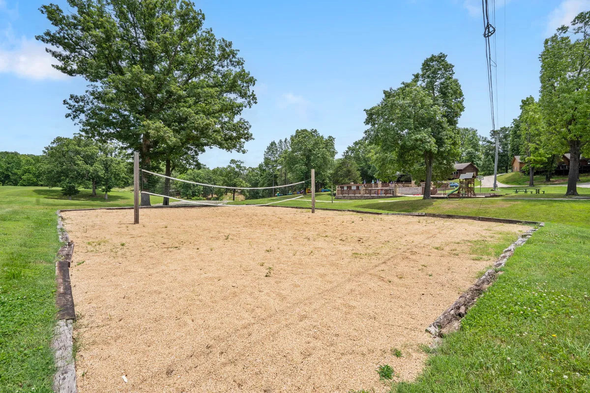 A cheerful sand volleyball court sits amidst green grass and trees, with cabins visible in the distance. The space is bustling with activity as families and friends enjoy friendly volleyball matches and picnics in a lively setting.