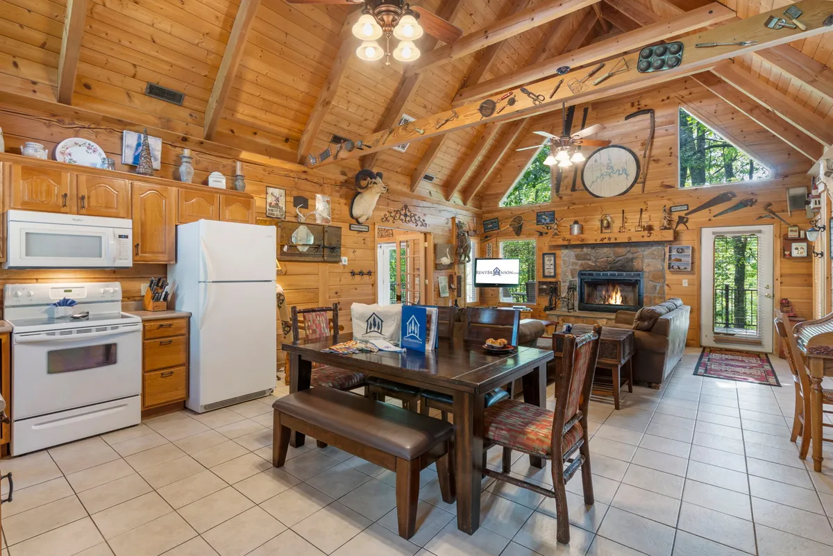 A warm, open-concept interior focusing on the kitchen island, dining area, and the fireplace beyond. Expansive windows illuminate the space, and the kitchen is clearly equipped for group meal prep and socializing, embodying the 'hub' where guests gather.