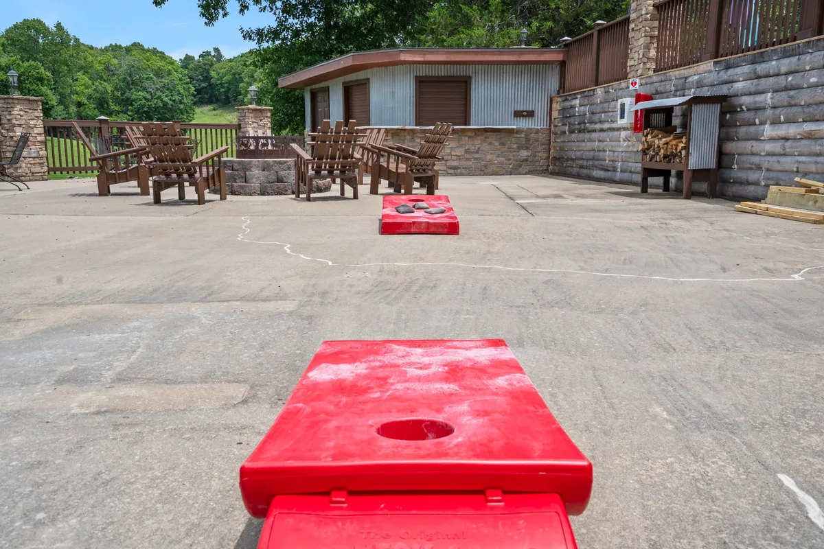 This image shows a lively outdoor area with red cornhole boards, wooden chairs circling a fire pit, a stone wall backdrop, and a cozy cabin-like structure. It evokes scenes of playful competition, shared laughter, and evenings spent around the fire.